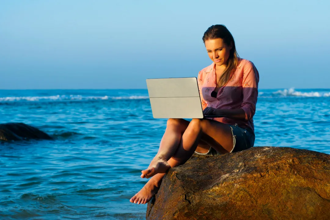 Mulher trabalhando com notebook na praia, simbolizando liberdade financeira